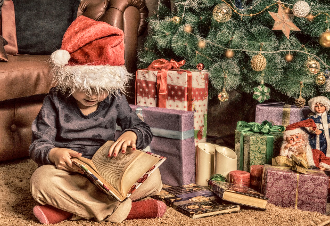 A photo by Aleksa_86, via Pixabay is of a Santa-hat wearing child sitting cross-legged by the Christmas tree opening a book.