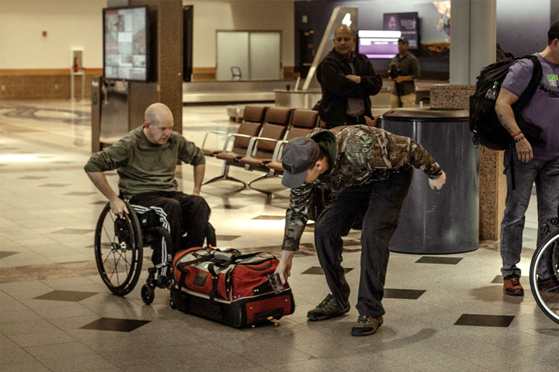 A man in a wheelchair is getting help from a fellow soldier in moving his duffle bag.