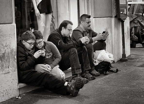Black-and-white photo of two boys and two men sitting on a step outside playing with their cellphones