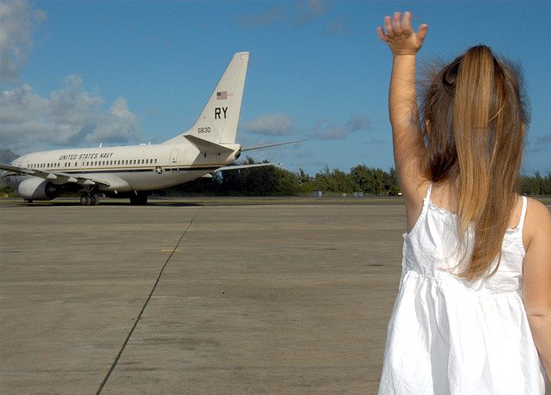 A young girl in a white sundress with a long ponytail waves goodbye to a US Navy plane