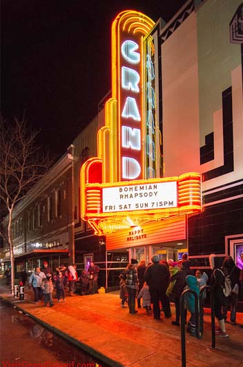A night scene with a brightly vertical sign proclaiming the movie Bohemian Rhapsody with people lining up outside of the historic Grand Theatre during Railside Christmas.