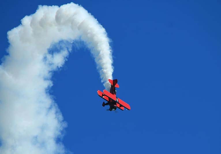 Against a bright blue sky, a red biplane flies straight down leaving a trail of smoke behind him.