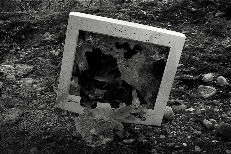 Black-and-white photo of a computer monitor with a bashed-in screen sitting in the woods