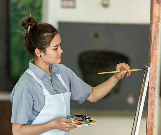 Profile view of a woman with her dark hair up, wearing a smock, and holding a palette of paints is painting on an easel