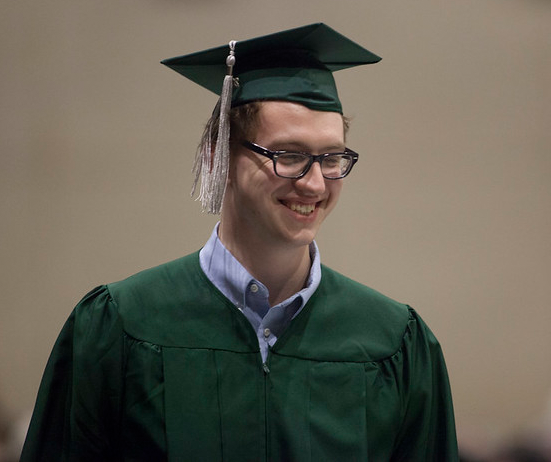 A lone man in a green gown wearing a green mortarboard with a pale creamy rose tassel dangling