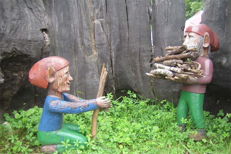 Two colorfully painted wooden gnomes in front of a huge treewith the one on the right kneeling in the grassin appeasement and the other standing with an armload of wood.