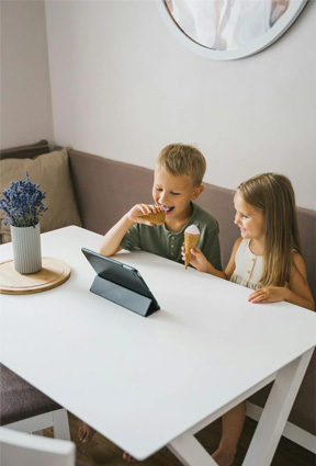 Two children are holding ice cream cones and sitting at a table with a tablet propped open on it.