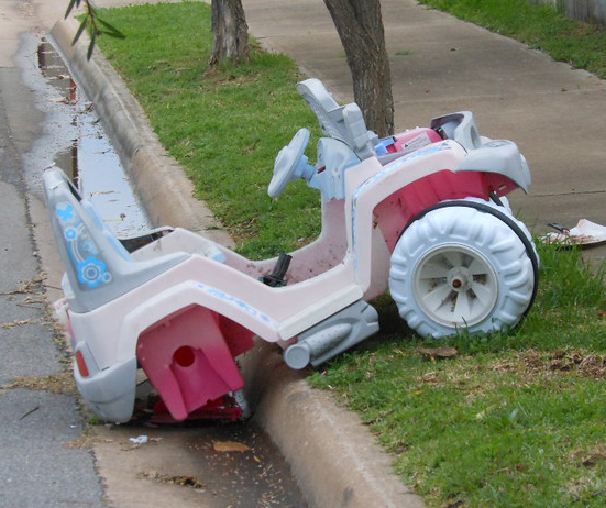 A plastic car in pastels is canted up against the curb with a broken wheel