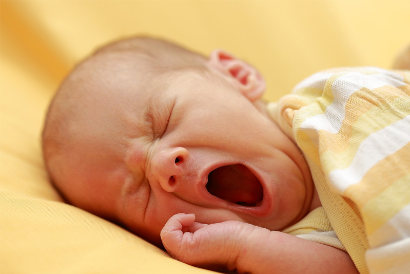 A baby yawning and wearing a yellow and white striped shirt while lying on a yellow sheet.