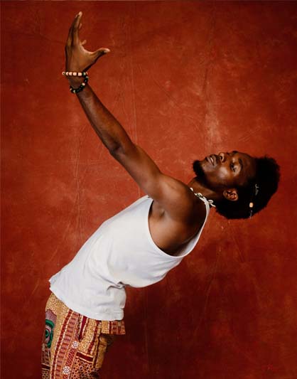 A bearded man with an Afro in a white tank top and print shorts is in profile facing left and bending backwards with his left arm up, his hand open yet curved wearing two bracelets against a deep orange-red background.