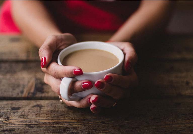 Close-up of a woman's hands holding a cup of coffee in a white cup above a rough wood table.