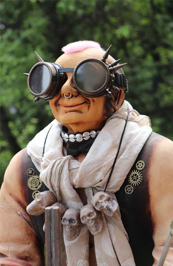 Man with a strip of fuzzy pink hair on his head, spiked goggles, a nose ring, a silver disk dog collar, a sack tied around his neck and brown paint on his upper body mimicking a vest with brass gears glued to his skin.