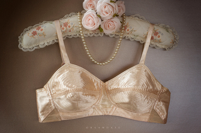 champagne rayon satin bra hanging from an elaborate hanger padded with a rose print, edged in lace with a cluster of roses at the base of the hook and pearls draped around the hook