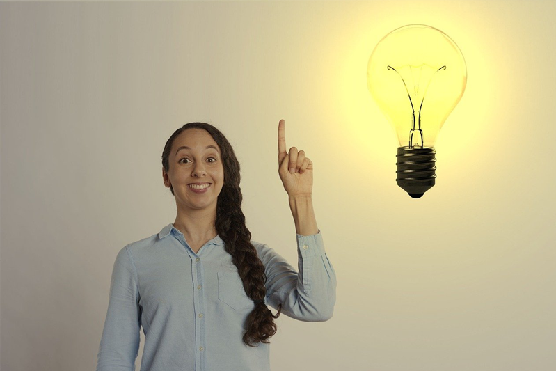 Smiling woman with long braid over one shoulder is holding up her left hand with her index finger pointing up. A light bulb is lit up on the right.