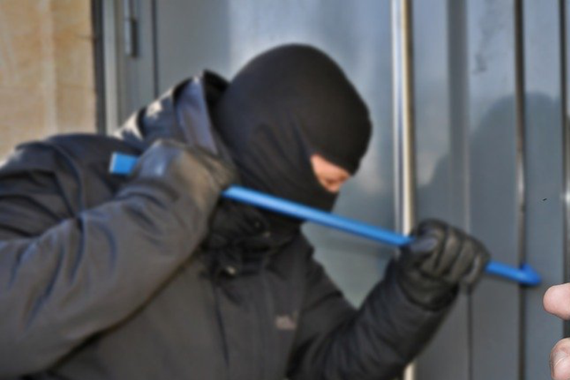 A man wearing a black hood, jacket and gloves uses a blue pry bar to prise open a sliding glass door.