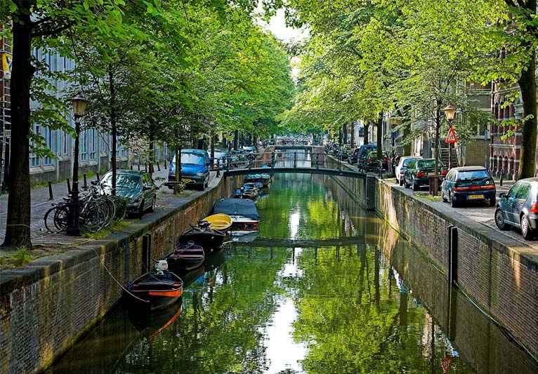 A straight-on view of a brick-lined canal with boats moored on the left and parked cars and bicycles, roads, and houses on either side.