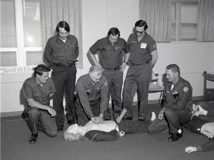 A black-and-white photo of five men surrounding another man performing CPR on a dummy.