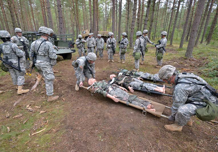 A group of soldiers in the woods in camouflage uniforms prepare to evacuate with three wounded soldiers on stretchers.