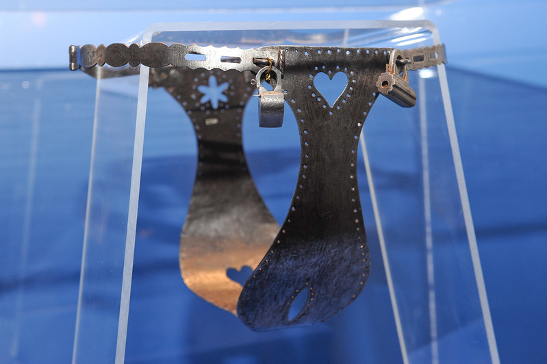 A metal chastity belt with two locks inside a plexiglass case against a blue background.