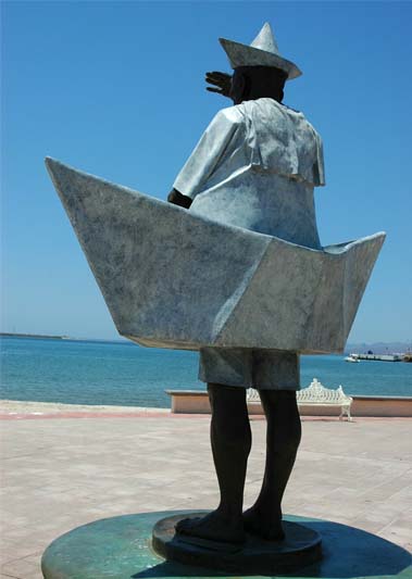 Bronze statue of a man in a wedge hat and caped jacket in an adult-sized paper boat, looking out over the sea with a clear blue sky over the boardwalk in La Paz, Baja California Sur, Mexico