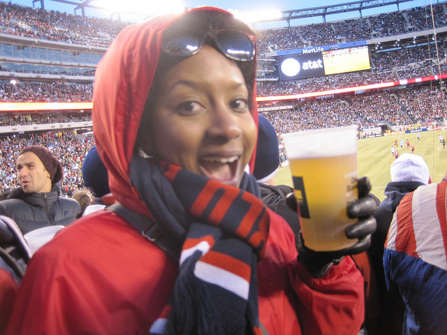 A woman in a jacket and hat, wrapped up in a scarf holds a plastic cup of beer at a stadium