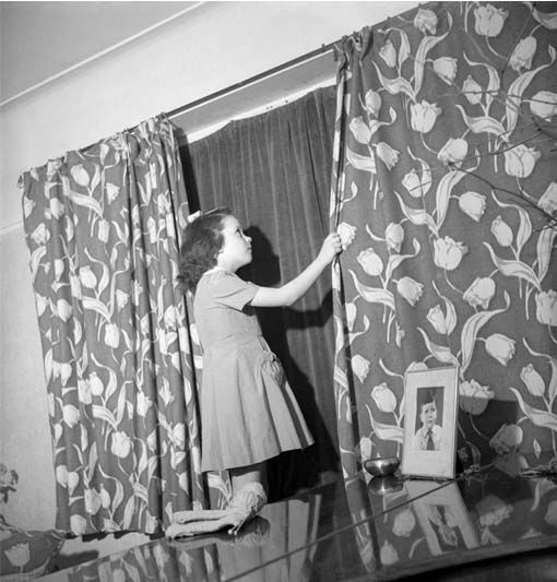 A black-and-white photo of a young girl in a dress pulling curtains across a window.