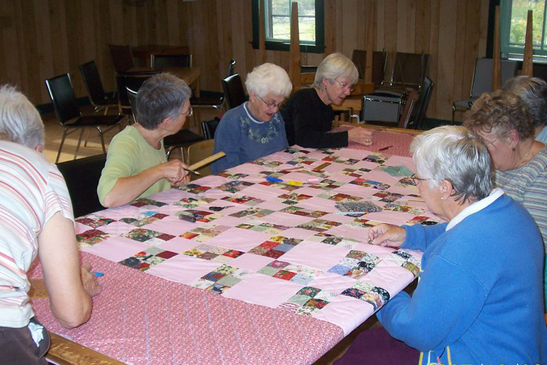 Seven older ladies sitting around a pink patchwork quilt quilting it in a dark wood-paneled room