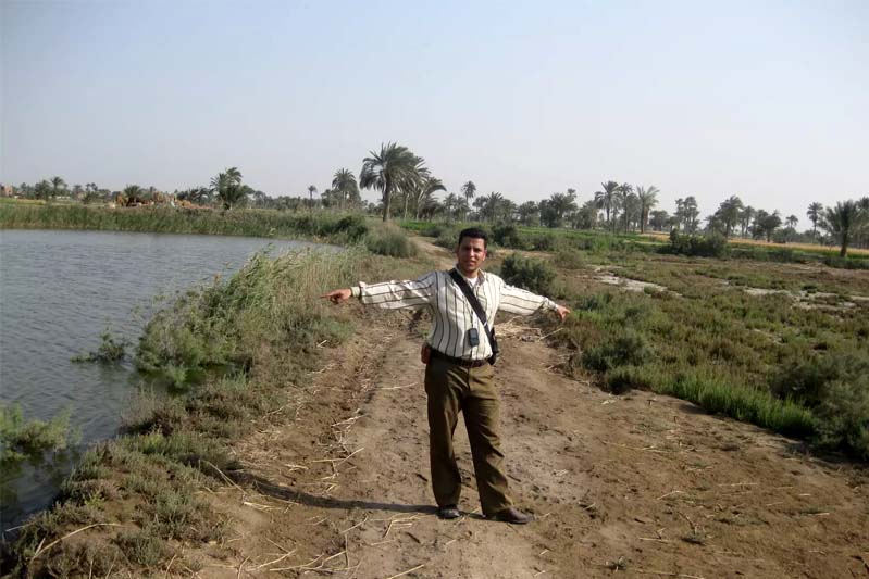 Man standing between a fish pond and a field.