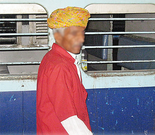 Man in a red shirt over a white one and a yellow turban stands next to a blue-and-white train car