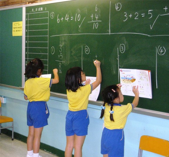 Three schoolgirls with black hair and wearing yellow tops and royal blue shorts are writing on the chalkboard