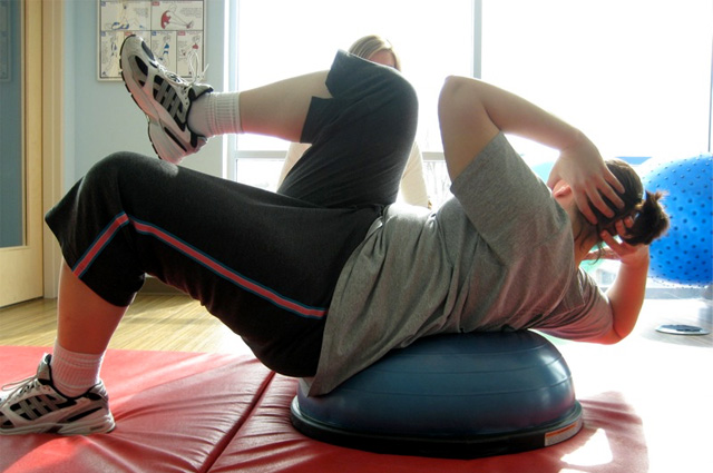 A woman in yoga pants and a gray T has one leg bent at the knee and raised over the other knee while twisting her torso in the opposite direction with her hands behind her head while propped up on soft half-dome base.