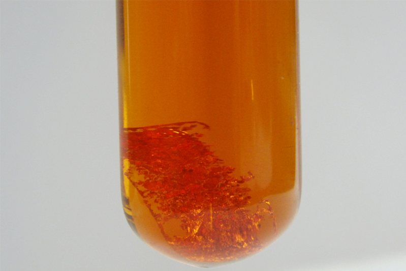 Close-up of the bottom of a test tube with a chunk of gold dissolving in a yellow-orange fuming liquid.