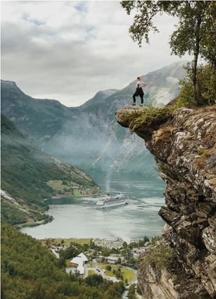 Soft, grayed colors depict the cruise ship steaming down the river with high mountains surrounding it. A woman stands in a dominant pose on an outcropping of rock above a village below her