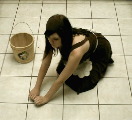 Woman in black tank top and pants on her hands knees scrubbing a tiled floor