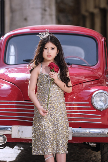 A girl standing in front of a vintage red car and wearing a tiara and glittery dress while holding a long-stemmed red rose