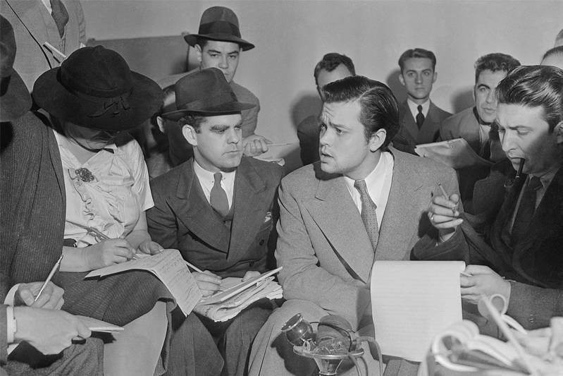 A profile of Orson Welles' face surrounded by reporters in a black-and-white photo