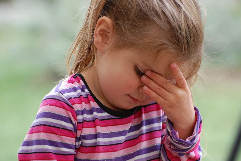 Cute little girl with blonde hair wearing a striped T-shirt has her head bowed and the fingers of one hand pinching the bridge of her nose.