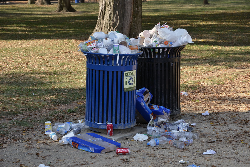 A close-up of a blue and a black recycle bin is overflowing with trash, some of which is on the ground.