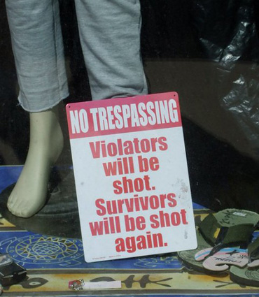 The lower half of a window display showing a woman's calves in capris and flip-flops on the lower right with a huge sign stating that there will be no trespassing. Violators will be shot. Survivors will be shot again.