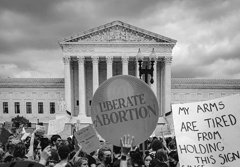 Black-and-white photo of an abortion protest. The sign on the left says Liberate Abortion while the sign on the right says My Arms are Tired From Holding This Sign.