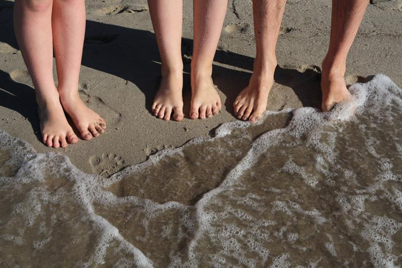 Three pairs of bare feet standing on the sand as a wave washes over them.