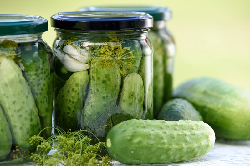 Three jars of pickles and some fresh cucumbers on the table.