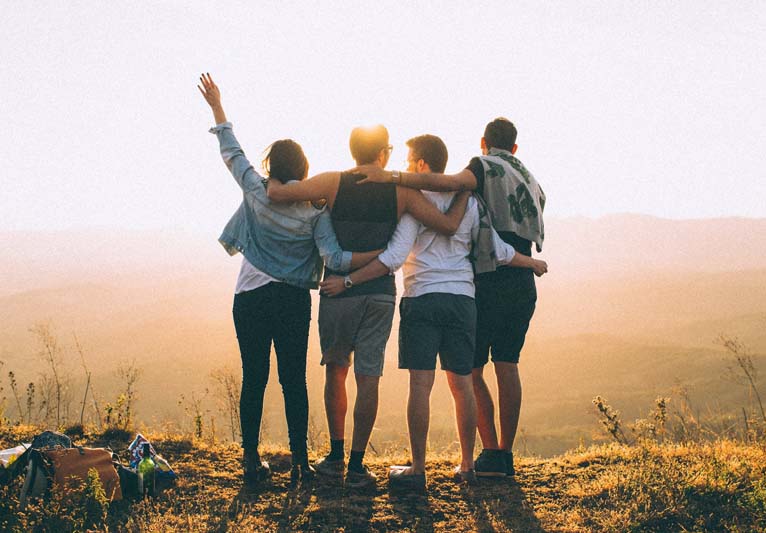 Four friends, standing arm in arm with their backs to us on top of a mountain looking out over a beautiful sunrise.