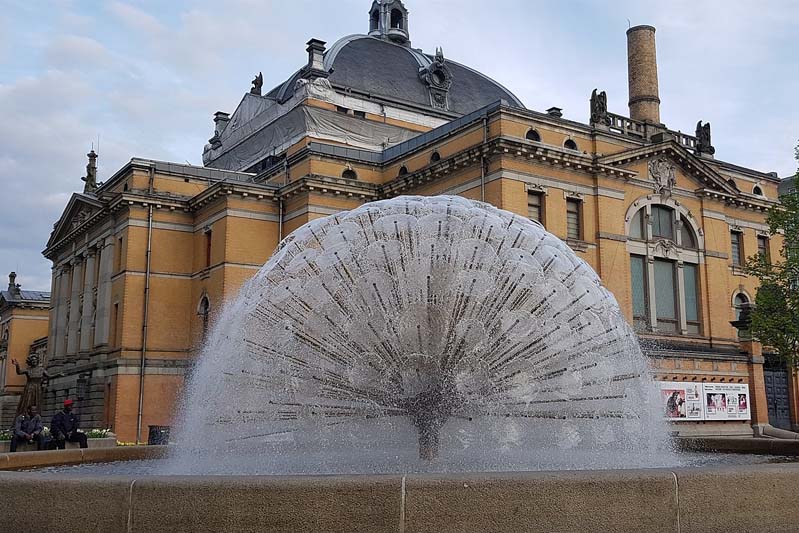 A large spiky fountain in front of a golden building forms a dome of water.