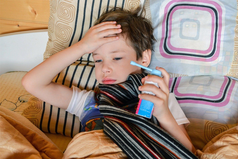 A young boy in a T-shirt lying against a bank of pillows holding an inhaler, a dark-striped scarf wrapped around his neck with one hand against his forehead
