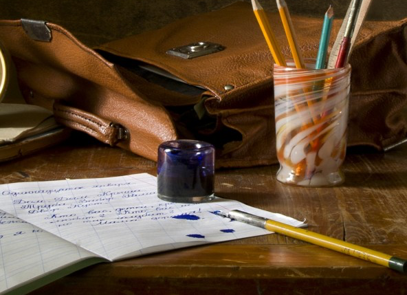 A still life of a journal, a stack ofbooks, an old-fashioned alarm clock, a leather satchel, a handblown glass of pens, a blue jar of ink, and a yellow ink pen on a scarred desk