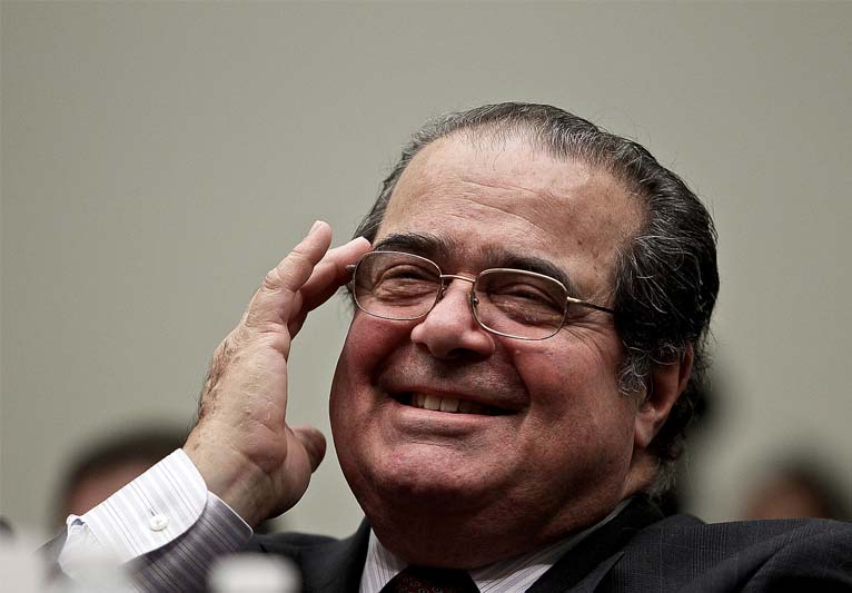 A close-up of Scalia's grinning face as he pushes up his glasses with his middle finger.