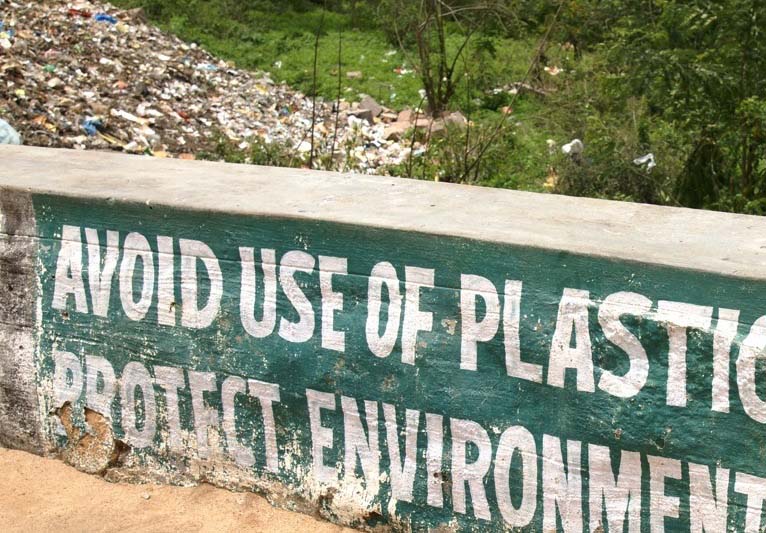 Behind a sign saying Avoid Use of Plastic Protect Environment is a huge mound of garbage.