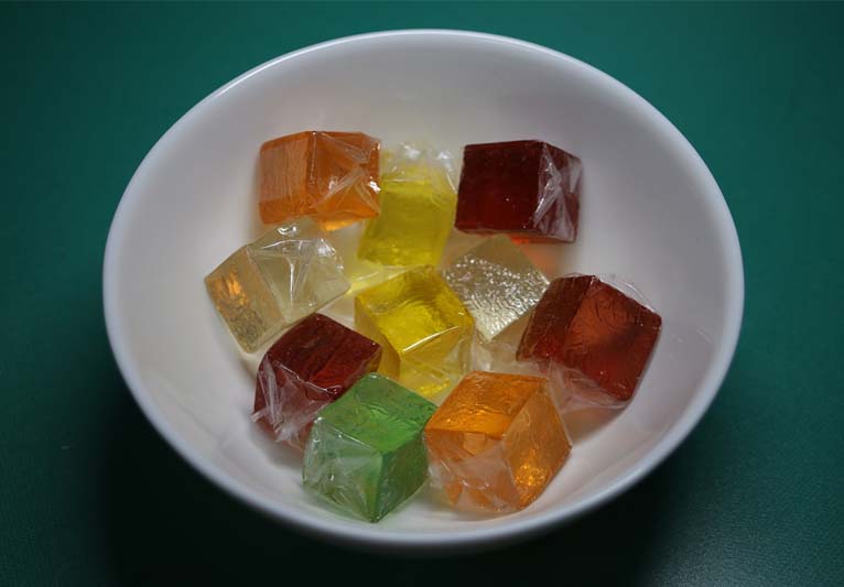Against a dark green background, a white bowl holds a variety of colored cellophane-wrapped jellies.