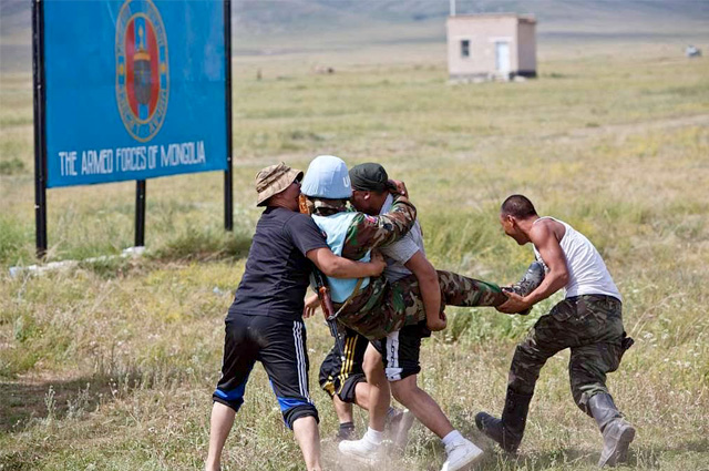 With a huge field as a backdrop with a signboard and a small building in the background, three men are carrying another man.